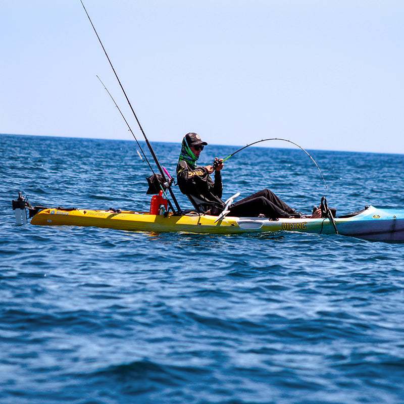 Angler in a yellow and blue Viking fishing kayak with a Bixpy electric motor, reeling in a catch on open water under a clear sky.
