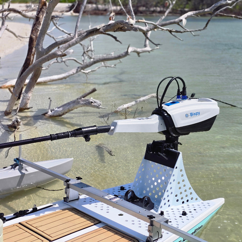 White Bixpy electric outboard motor mounted on a perforated stand with a tiller handle on a paddleboard, in very shallow clear water with sandy bottom and trees.