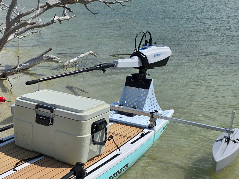 White Bixpy electric outboard motor mounted on a perforated stand with a tiller handle on a paddleboard, next to a cooler in shallow water with tree branches.