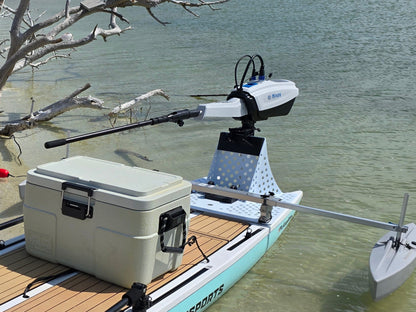 White Bixpy electric outboard motor mounted on a perforated stand with a tiller handle on a paddleboard, next to a cooler in shallow water with tree branches.