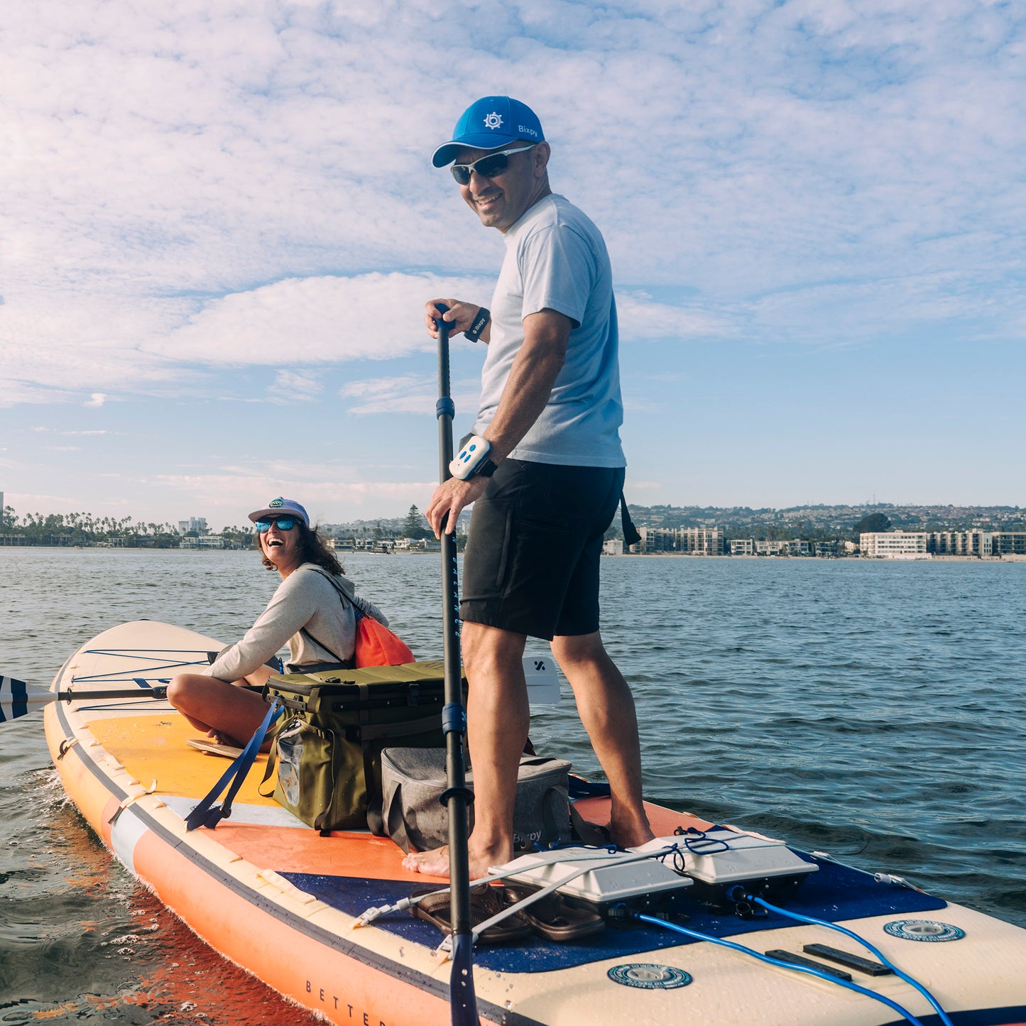 Two people on a Bixpy-powered stand-up paddleboard with two mounted batteries, a motor, and a bag, on a bay with a distant shoreline.