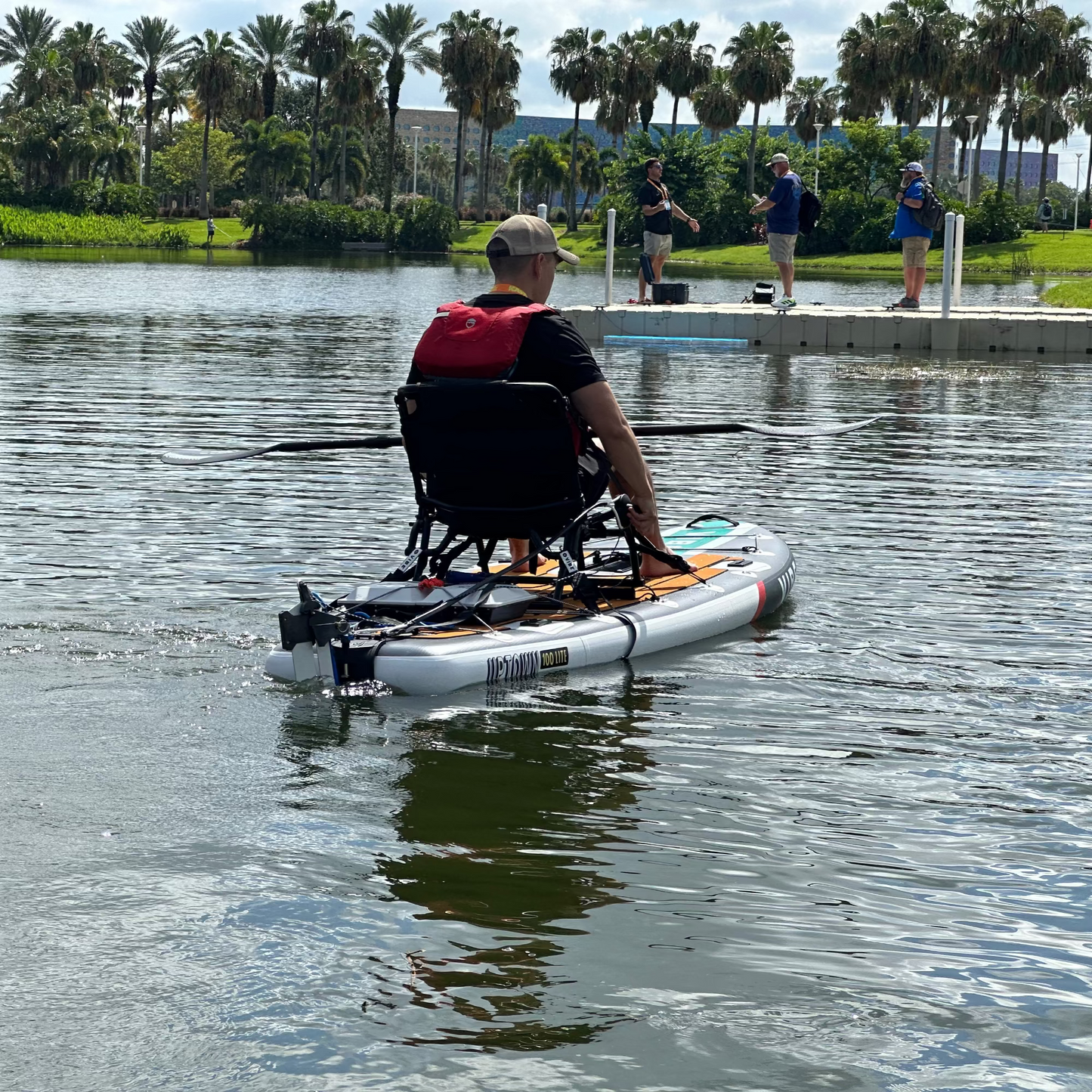 Person in a white paddleboard with a seat, propelled by a Bixpy motor, paddling on a lake with a fishing dock and palm trees in the background.