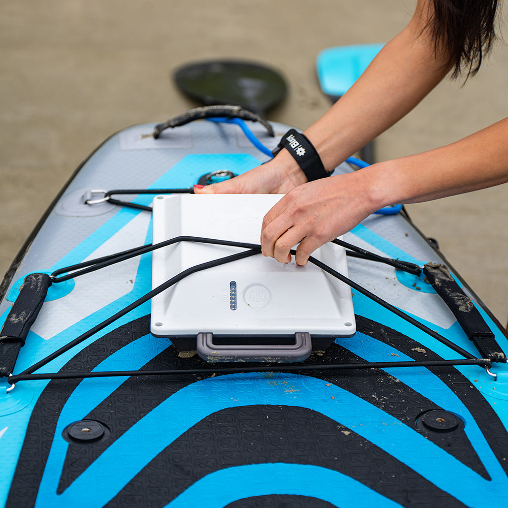 A person's hands securing a Bixpy battery to a blue and grey inflatable paddleboard with bungee cords.