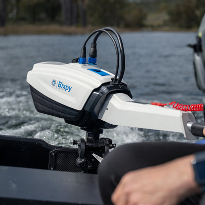 Close-up of a white and black Bixpy electric motor battery pack mounted vertically on a black bracket attached to a blue inflatable tube, with cables connected and water in background.