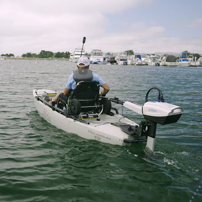 A person in a Hobie Pro Angler 12 kayak equipped with a wheelchair seat paddles away from the camera on calm water. Mounted on the back of the kayak is a Bixpy N-3 outboard motor, installed using the Bixpy Power Pole to Transom Conversion Adapter. In the background, boats and yachts are docked at a marina under a partly cloudy sky.