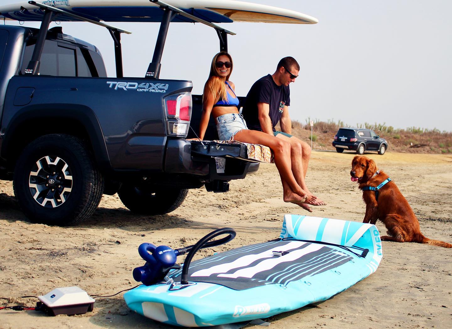 Man and woman sit on a truck tailgate with dog nearby and watercraft sitting on the sand with a Bixpy Motor